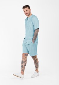 Uomo con tatuaggi su braccia e gambe che cammina, indossando una camicia azzurra a maniche corte con bottoni, pantaloni corti coordinati e sneakers bianche su sfondo bianco.