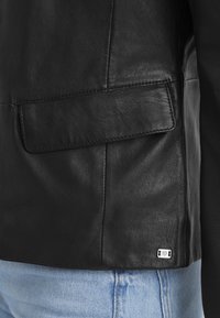 Černá kožená bunda se hladkým povrchem, se čtvercovou kapsou a malou bílou značkovou nálepkou ve spodní části, nošená přes světle modré džíny.