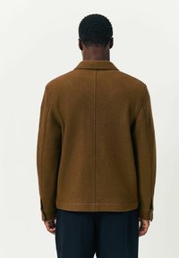 Ein Mann von hinten zu sehen, der eine braune Wolljacke mit Nahtdetails und dunkelblaue Hosen vor einem schlichten weißen Hintergrund trägt.