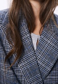 Blazer de mezcla de lana con un patrón de pata de gallo en azul y gris, solapas con muesca y un acabado texturizado. Se ve parcialmente una prenda superior gris debajo.