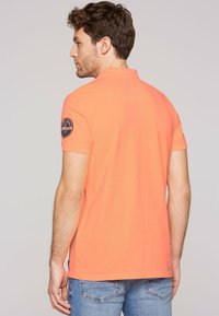 Polo naranja con un acabado texturizado, mangas cortas, detalle en el hombro y un sutil logo en la parte posterior. Combinado con jeans azules.