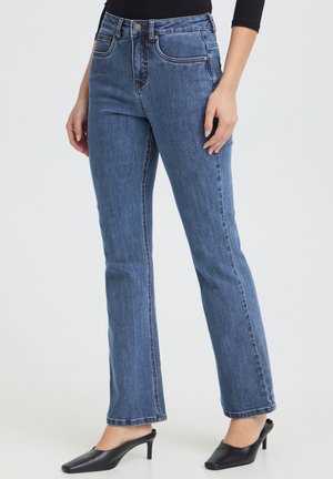Femme portant un jean bleu droit associé à des chaussures noires à petit talon sans lacets, debout devant un fond clair uni.