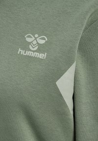 Tissu vert texturé avec le logo blanc Hummel, comprenant une abeille stylisée au-dessus du nom de la marque et un motif partiel en chevron blanc.
