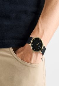 Mustriga must kell, millel on kuldsed detailid ja must nahast rihm, käe peal, mis on asetatud beežide pükste taskusse.