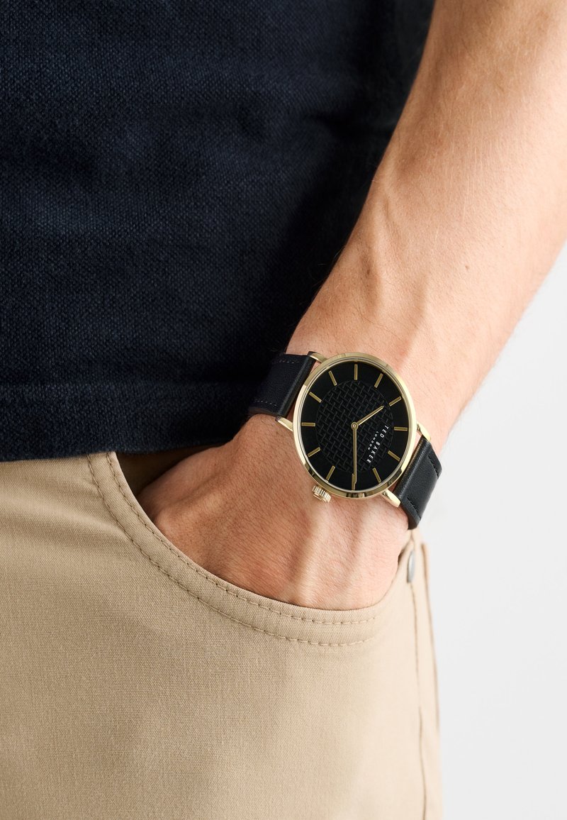 Mustriga must kell, millel on kuldsed detailid ja must nahast rihm, käe peal, mis on asetatud beežide pükste taskusse.