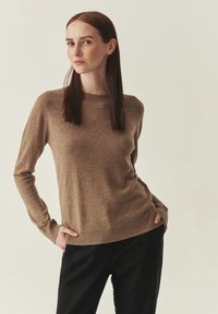 Pull en maille brun clair avec un col rond et des manches longues, présentant un ourlet et des poignets côtelés. Associé à un pantalon noir.