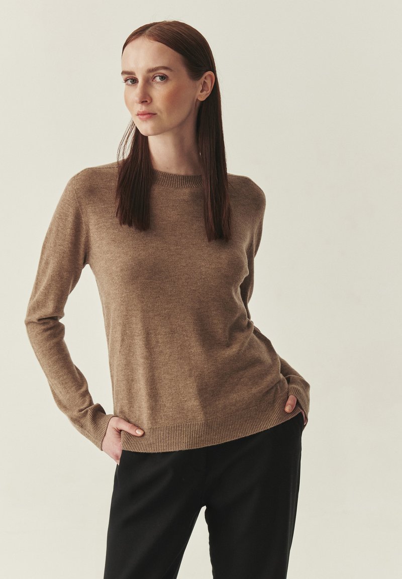 Pull en maille brun clair avec un col rond et des manches longues, présentant un ourlet et des poignets côtelés. Associé à un pantalon noir.