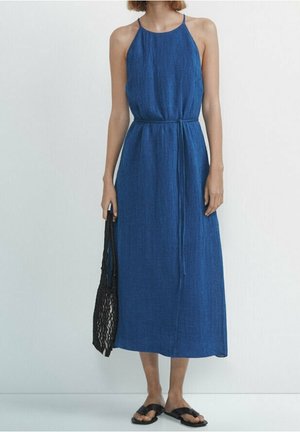Vestito azzurro con collo a fascia realizzato in tessuto leggero, caratterizzato da una vita increspata, una silhouette fluente e una lunghezza fino a metà polpaccio. Indossato con sandali bassi.