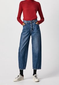 Person som bär högmidjade vida blå jeans, svarta strumpor, vita sneakers och en röd ribbad långärmad topp med händerna i fickorna.