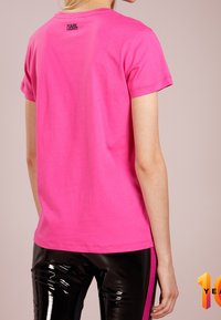 Rosa Baumwoll-T-Shirt mit kurzen Ärmeln, versehen mit einem kleinen schwarzen Logo auf der Rückseite. Getragen mit glänzenden schwarzen Hosen, die den Textur-Kontrast betonen.