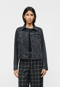 Γυναίκα με μαύρο τζιν μπουφάν με τρουκς πάνω από μαύρη μπλούζα και καρό παντελόνι σε ασπρόμαυρο, στέκεται μπροστά σε απλό φόντο.