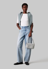 Jeans baggy blu chiaro, t-shirt bianca aderente e un gilet senza maniche grigio chiaro in maglia. Tiene una piccola borsa a tracolla grigia. Scarpe nere.