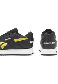 Čierne tenisky s žltými akcentmi, s hladkým koženým zvrškom, bielou medzipodrážkou a štruktúrovanou gumovou podrážkou. Logo "Reebok" viditeľne umiestnené.