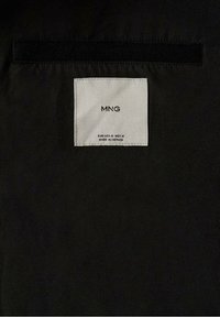 Étiquette textile noire avec des coutures blanches, portant "MNG" et des instructions d'entretien. Attachée à une surface de tissu noir lisse.