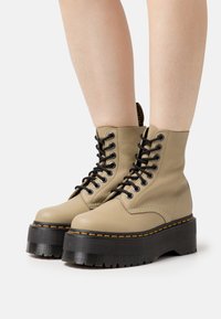 Stivali da combattimento in pelle beige con zeppa e suole spesse nere, lacci neri e cuciture gialle, indossati su gambe nude su uno sfondo bianco.
