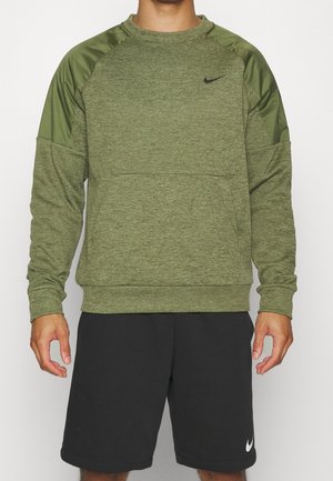 Man draagt een olijfgroene Nike trui met lange mouwen en patches op de schouders en zwarte Nike shorts, staand tegen een effen achtergrond.
