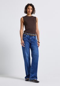 Frau in einem dunkelbraunen ärmellosen Top, blauen weit geschnittenen Jeans mit einem Gürtel im Leopardenmuster und schwarzen spitzen Ballerinas vor weißem Hintergrund.