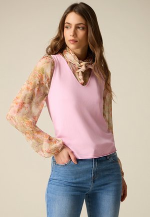 Femme aux longs cheveux bruns portant un haut rose sans manches sur un chemisier fleuri transparent noué au cou, et un jean bleu, regardant vers la gauche.