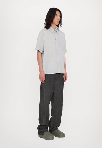 Chemise à manches courtes gris clair à boutons avec des détails de couture subtils, associée à un pantalon large gris foncé et des chaussures vertes à enfiler.