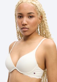 Γυναίκα με ανοιχτόχρωμο δέρμα και μακριά ξανθά κοτσίδια, που φοράει λευκό επενδεδυμένο σουτιέν με λεπτούς ιμάντες και δαντελένια διακόσμηση, με ουδέτερη έκφραση, απλό φόντο.