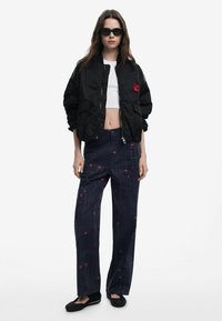 Čierna bomber bunda s červenou výšivkou, biela skrátená blúzka, námornícke patterned nohavice s červenými kvetinovými akcentmi a čierne baleríny. Slnečné okuliare dokončujú vzhľad.