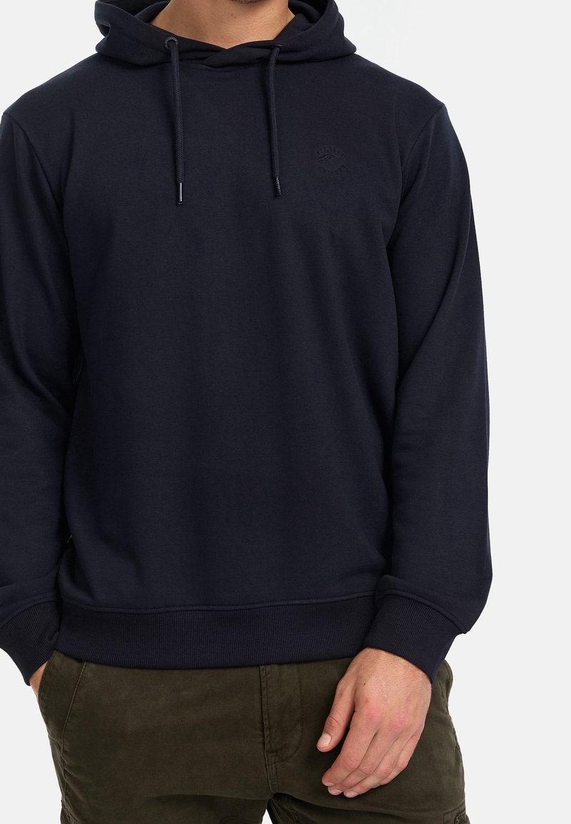 Homme portant un sweat à capuche noir avec cordons et un pantalon vert olive sur un fond uni.