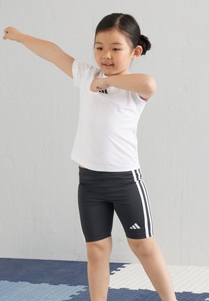 Camiseta atlética de manga corta blanca con logo negro, combinada con pantalones cortos negros que tienen rayas laterales blancas. El fondo es una pared gris texturizada.