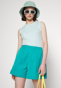 Top sem mangas azul claro combinado com calções de elástico teal. Os acessórios incluem um chapéu de balde verde menta e óculos de sol quadrados. Alça da bolsa amarela.