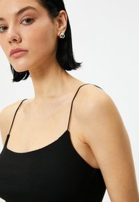 Zwarte tanktop met dunne bandjes, voorzien van een strakke pasvorm en geribde textuur. Model draagt grote zilveren stud oorbellen. Minimalistisch ontwerp.