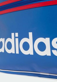 Modra športna torba za prenašanje z veliko belo nalepko, rdečimi zadrgami in gladkim sintetičnim materialom. Vključuje preprost in funkcionalen dizajn.