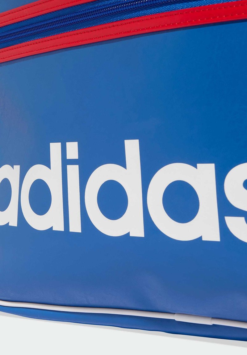 Borsa sportiva atletica blu con un grande logo bianco, dettagli della cerniera rossa e materiale sintetico liscio. Include un design semplice e funzionale.