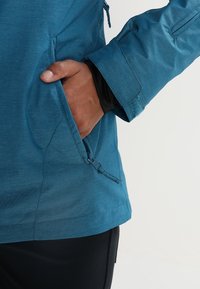 Teejake mit sichtbarem Handtaschenfach. Der Stoff ist glatt mit einem leichten Glanz. Verfügt über einen Reißverschluss und ein tailliertes Manschetten-Design.