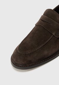 Mocasines de ante en marrón oscuro, con una textura suave, acentos cosidos y un diseño de punta redonda con un sutil detalle de corte.