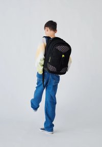Black backpack with checkered accents, durable fabric, padded back, and adjustable straps. Yellow zipper pull visible. Casual denim jeans worn.