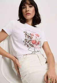 Biała bawełniana koszulka z grafika w kwiaty i w panterkę. Krótkie rękawy oraz okrągły dekolt. Połączona z jasnymi dżinsami.