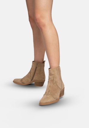 Stivaletti alla caviglia in camoscio color cammello con borchie decorative, cerniere laterali e tacchi bassi a blocco, indossati su gambe nude su sfondo semplice.