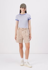 Shorts beiges avec un devant plissé, associés à un t-shirt à rayures bleues avec un col bleu, et une casquette verte. Baskets blanches avec des semelles crème.