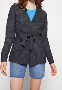 Mörkgrå stickad cardigan med knytmidja, öppen front och ribbad textur. Bärs över en blå topp och är ihop med jeansshorts.