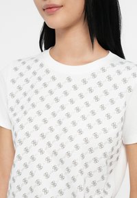 Hvit t-skjorte med et gjentatt grått mønster og et signaturlogo. Stoffet ser mykt ut med en standard rund hals og korte ermer.
