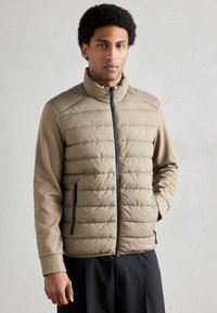 Doudoune beige avec fermeture éclair complète, devant matelassé et manches douces. Comprend des poches latérales avec fermetures éclair noires. Texture lisse et design structuré.