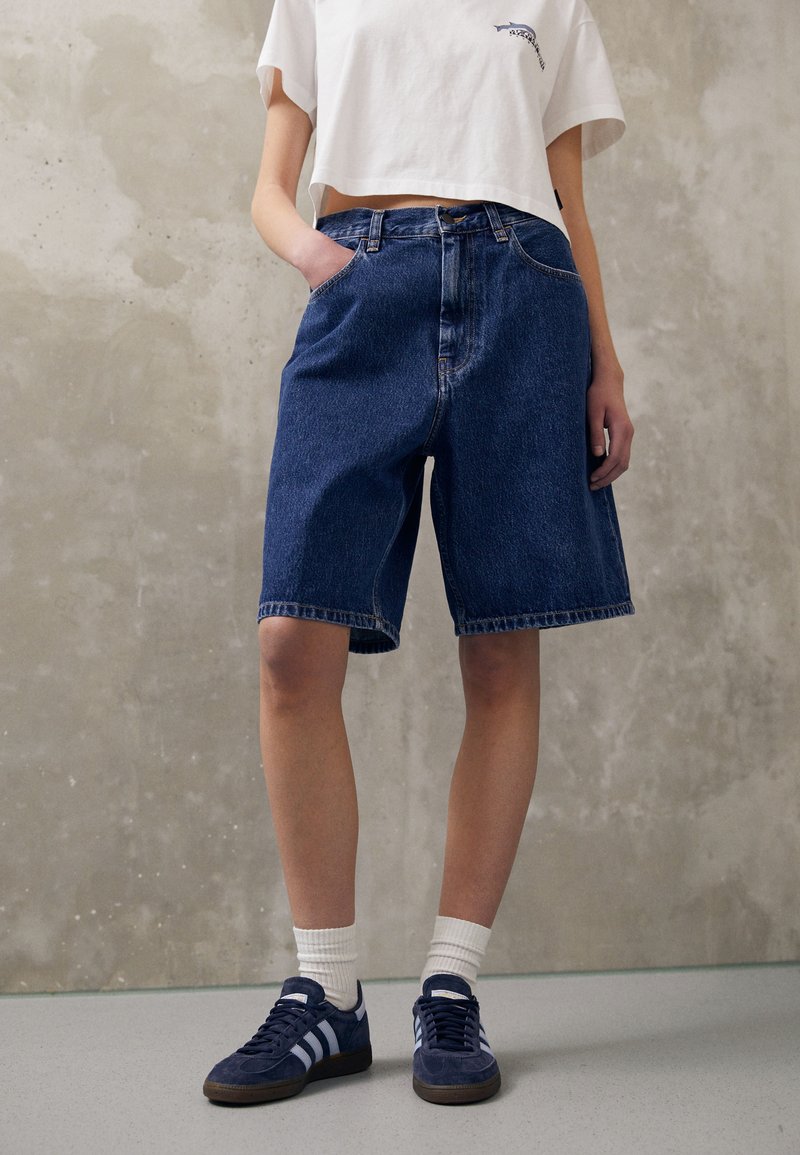Mørkeblå denimshorts med løs passform, med frontlommer og synlige sømmer. Kombinert med marineblå sneakers og hvite ankelsokker.