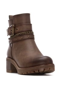 Botín de cuero marrón hasta el tobillo con un tacón grueso, que presenta una cadena decorativa y una correa con tachuelas. Suela de goma con un patrón texturizado.