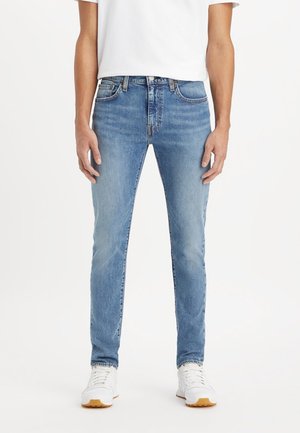 Person trägt hellblaue Slim-Fit-Jeans, weiße Sneakers und ein weißes kurzärmeliges Hemd und steht vor einem einfachen weißen Hintergrund.