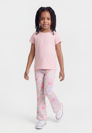 Young girl with braided hair wearing a pink t-shirt, floral patterned pants, and white sneakers, standing with one leg slightly raised.