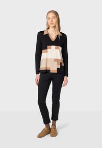 Pull en V noir avec un motif géométrique beige, crème et marron. Associé à un pantalon noir ajusté et des baskets marron.