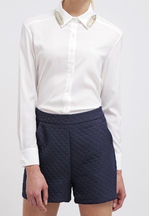 Vrouw draagt een wit overhemd met knopen met zilveren bladdetails op de kraag en marineblauwe doorgestikte shorts met hoge taille.