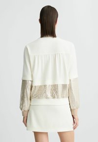 White athletic jacket with gold sequin accents on sleeves. Features a cropped design with a relaxed fit and ribbed cuffs.