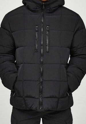Osoba ubrana w czarną pikowaną kurtkę puchową z kapturem i dwoma pionowymi zamkami na piersi, ręce w bocznych kieszeniach.