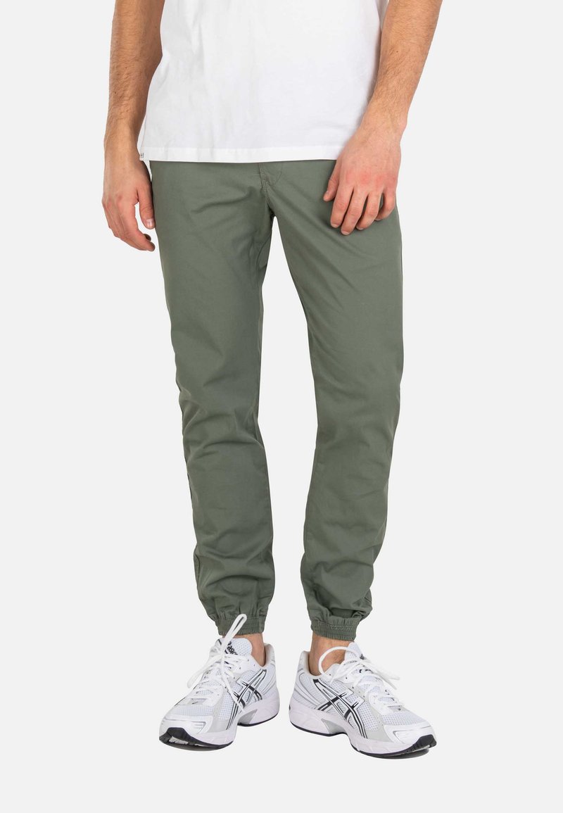 Hombre vistiendo pantalones jogger verde oliva y zapatillas deportivas blancas con detalles en negro, de pie sobre un fondo blanco.