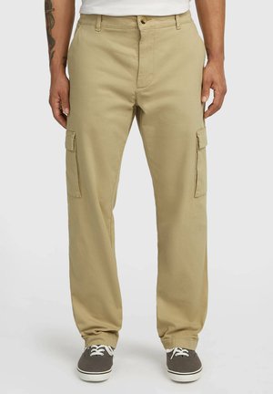 Man draagt beige cargobroek met zijzakken, wit T-shirt en donkergrijze vetersneakers, staand voor een effen achtergrond.